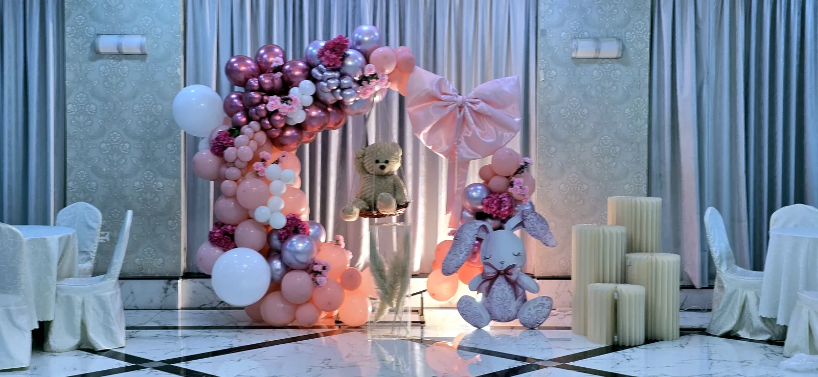 Arche de ballons roses et bordeaux avec un nœud, un ours en peluche et un lapin en peluche sur un sol en marbre