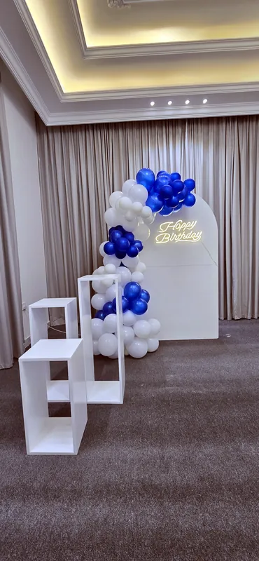 Гирлянда из белых и синих шаров у белой арки с золотой надписью Happy Birthday и кубами-подставками