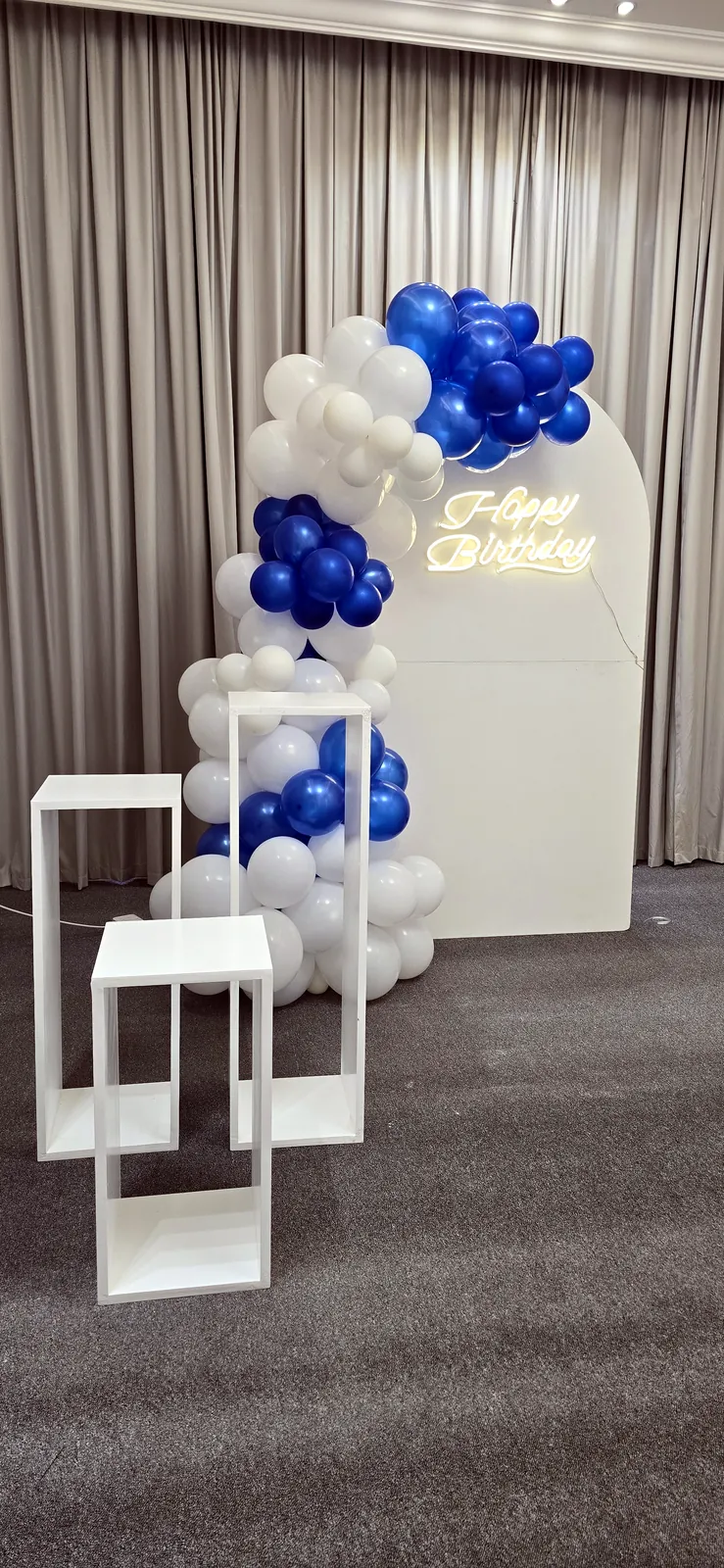 Guirlande de ballons blancs et bleus avec une inscription dorée Happy Birthday et des présentoirs cubes près de fenêtres à rideaux